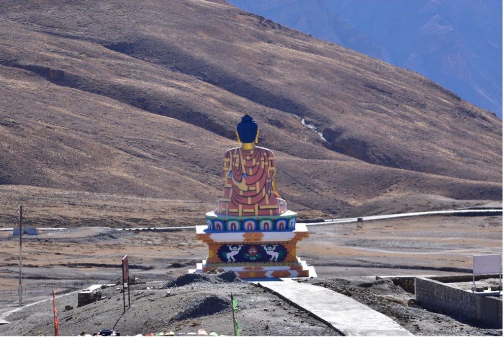 स्पीति घाटी में एक विशाल, रंगीन बुद्ध प्रतिमा एक चबूतरे पर विराजमान है, जिसके पृष्ठभूमि में एक विशाल पर्वतीय परिदृश्य दिखाई देता है।