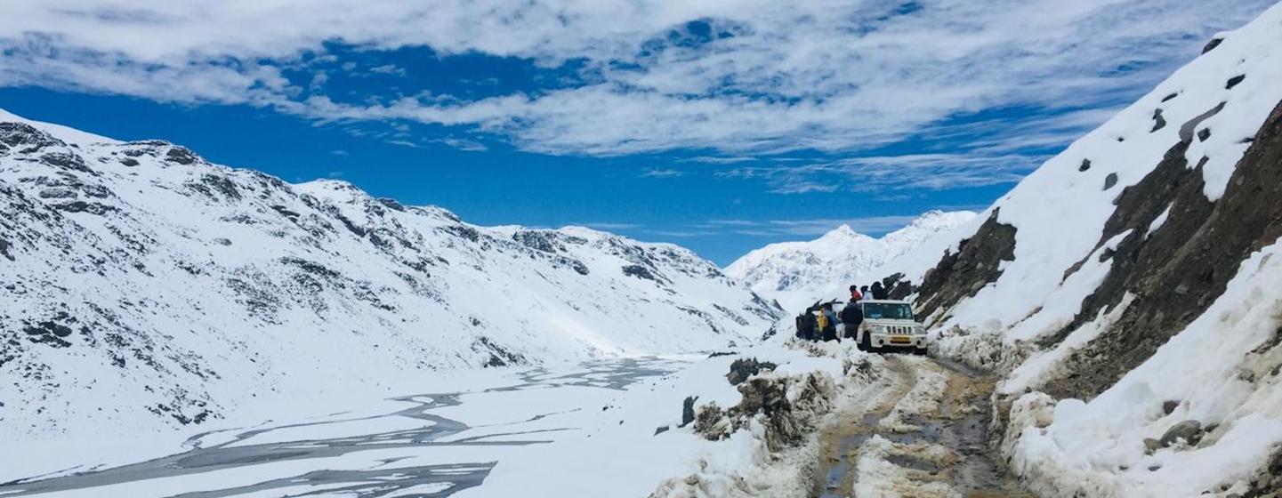 आंशिक रूप से बादलों से घिरे आसमान के नीचे हिमालय के एक पहाड़ी दर्रे से गुजरने वाली बर्फीली सड़क पर एक वाहन और पर्यटक।