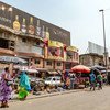 Vendeurs ambulants et clients au marché d'Adjamé à Abidjan, en Côte d'Ivoire. On déambule le long d'une rue pavée bordée d'étals proposant des marchandises sous des parasols colorés, avec des immeubles commerciaux en arrière-plan.