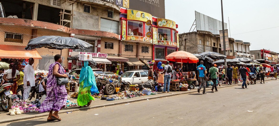 Vendeurs ambulants et clients au marché d'Adjamé à Abidjan, en Côte d'Ivoire. On déambule le long d'une rue pavée bordée d'étals proposant des marchandises sous des parasols colorés, avec des immeubles commerciaux en arrière-plan.