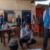 Naibu Katibu Mkuu UN Tom Fletcher, aliyevaa fulana ya bluu, amejilaza katika chumba katika kambi ya wakimbizi huko El Genina, Darfur, Sudan, huku akipewa taarifa na mfanyakazi mwingine wa misaada.