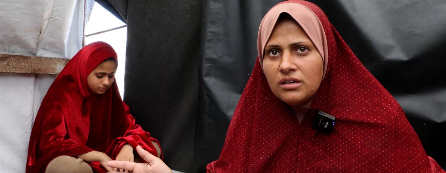 Uma mulher e uma menina, ambas vestidas de vermelho, estão sentadas dentro de uma tenda. A mulher conversa com alguém invisível, enquanto a menina senta-se atrás dela, olhando para baixo.