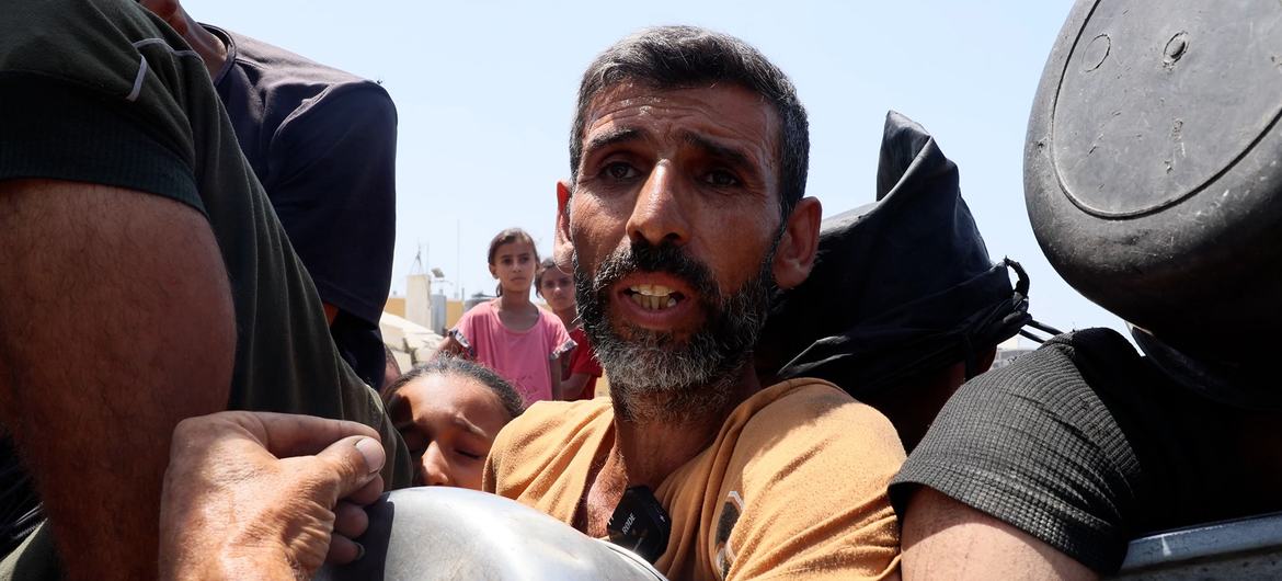 رجل نازح في غزة يحمل وعاءً معدنياً ويتحدث إلى الكاميرا، بينما يظهر أشخاص آخرون في الخلفية.