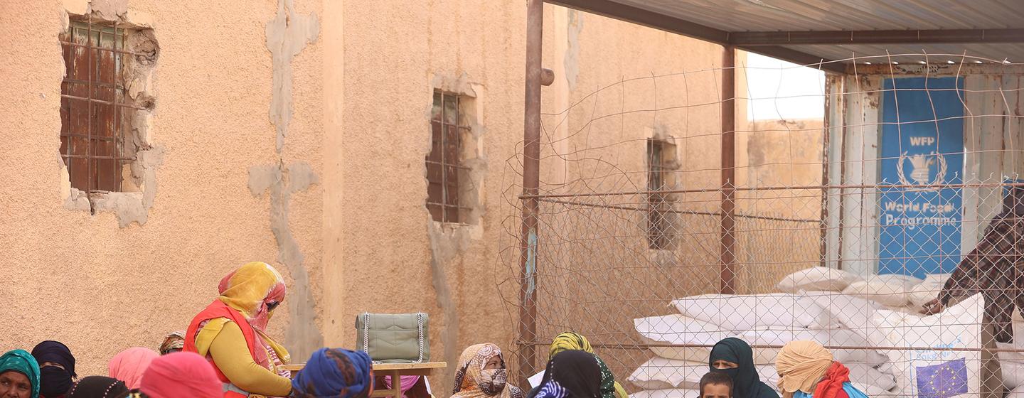 Mulheres e crianças recebem ajuda alimentar do Programa Mundial de Alimentos em um campo de refugiados na Argélia. Um trabalhador entrega um saco de grãos a uma mulher enquanto outras pessoas aguardam nas proximidades.