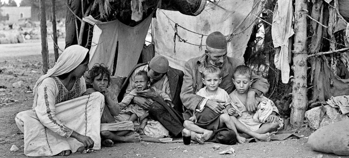 ایک سیاہ اور سفید تصویر میں ایک فلسطینی پناہ گزین خاندان کو خان ​​یونس میں اپنی عارضی پناہ گاہ کے باہر دکھایا گیا ہے۔ ایک خیمے نما ڈھانچے کے سامنے ایک عورت، دو مرد اور پانچ چھوٹے بچے زمین پر بیٹھے ہیں۔