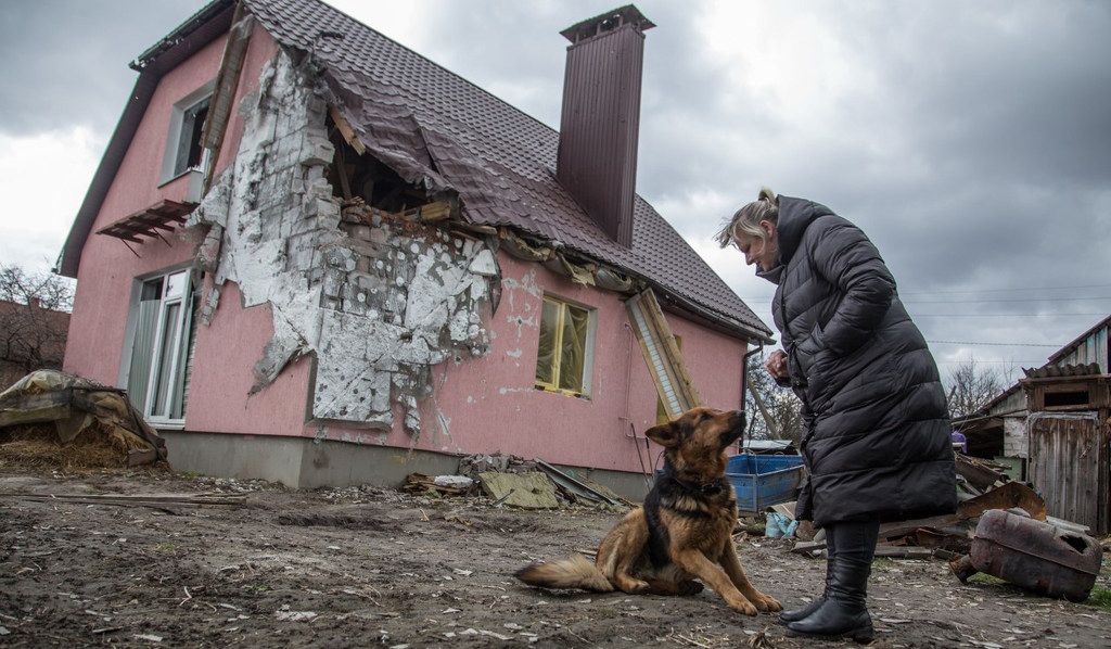 Une femme ukrainienne avec son chien se tient près d'une maison endommagée.