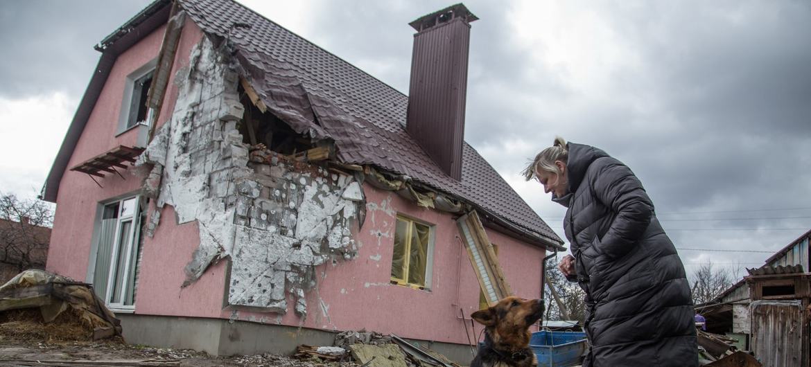 Mulher ucraniana com seu cachorro parada em frente a uma casa danificada