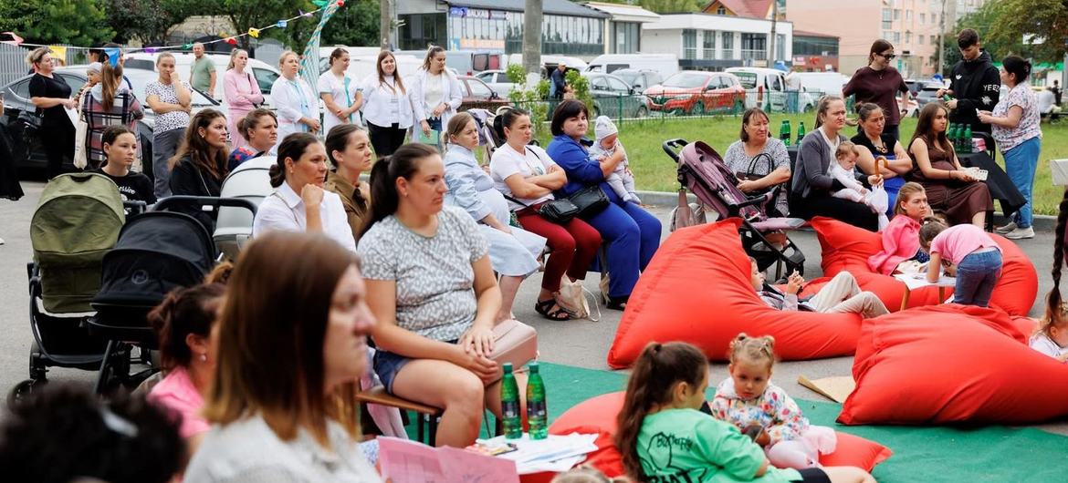 Lors d'un événement de santé publique en Moldavie, des mères et leurs enfants sont réunis sur des poufs et des chaises rouges, tandis que des soignants en blouse blanche se tiennent en arrière-plan.