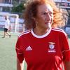 Пожилая женщина с кудрявыми рыжими волосами стоит на футбольном поле в красной футболке «Бенфики». На заднем плане видны другие игроки.