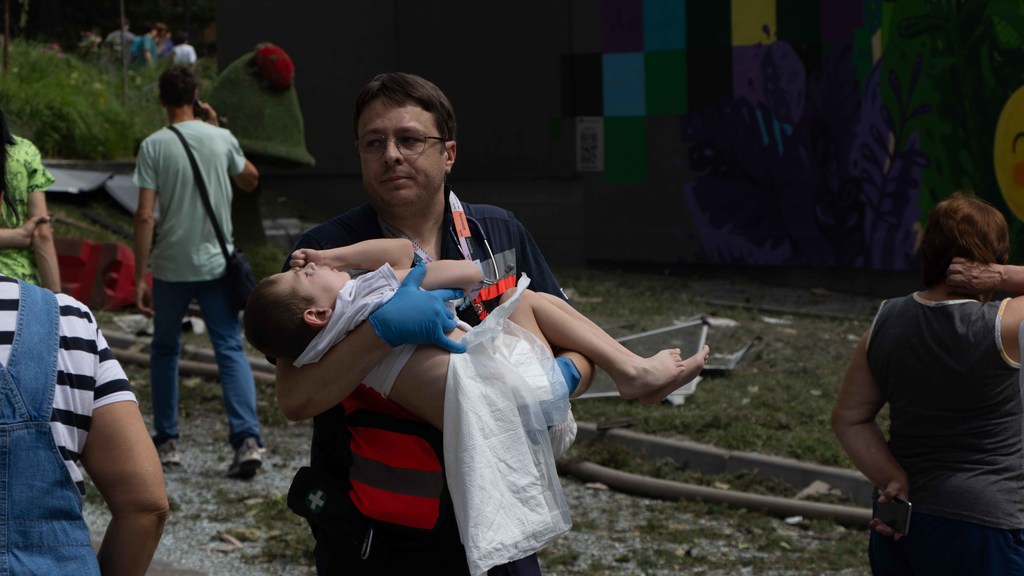 Un hombre con un chaleco rojo y negro y guantes azules lleva en brazos a un niño herido.