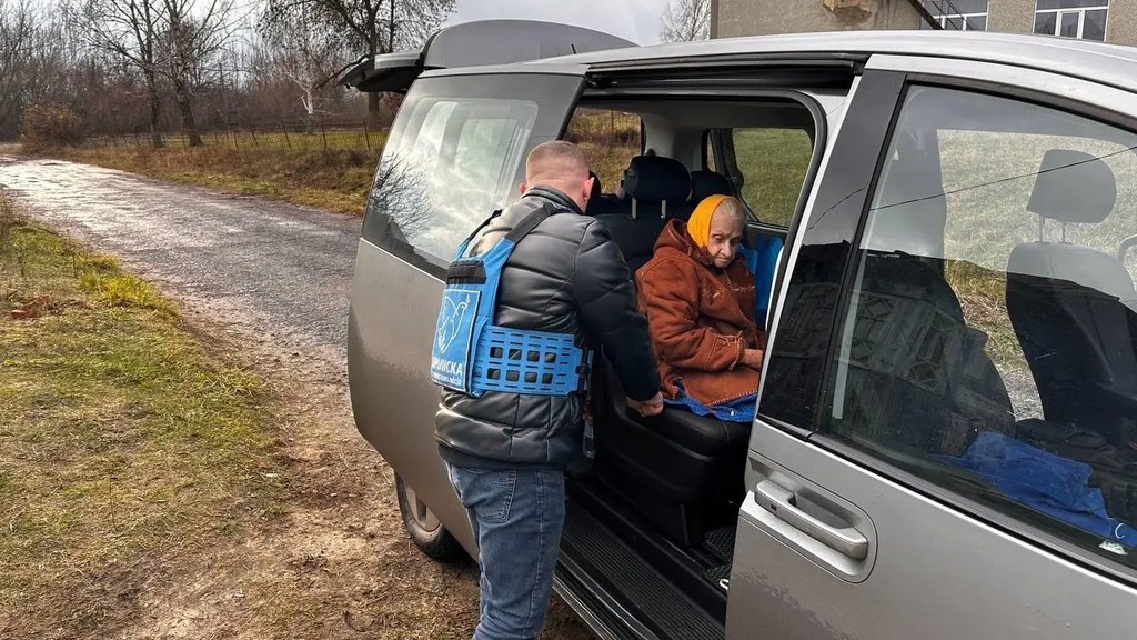 Мужчина в синем светоотражающем жилете помогает пожилой женщине в желтом платке сесть в заднюю часть серебристого фургона на сельской дороге.