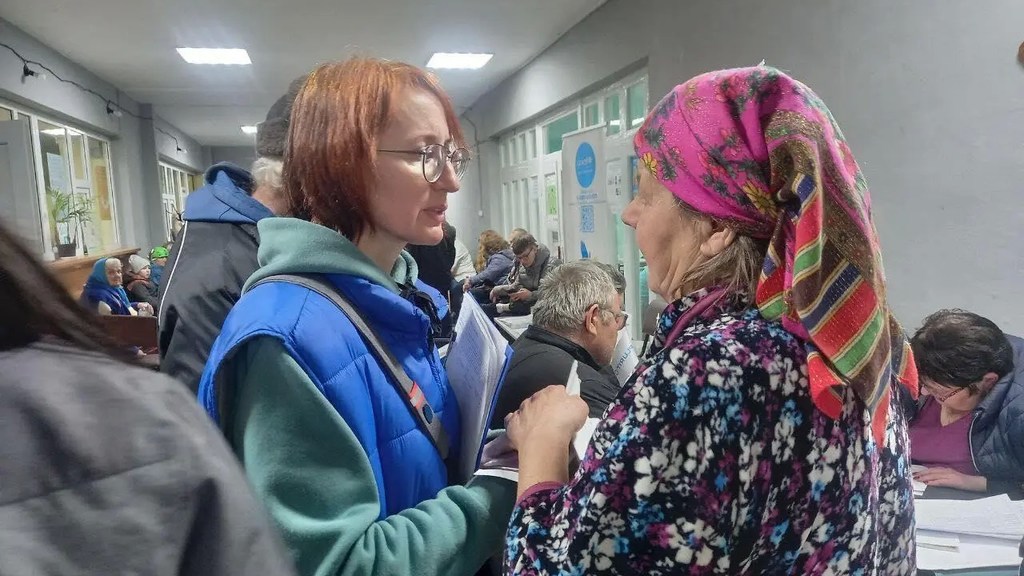 В общественном зале сотрудница гуманитарной организации с рыжими волосами и в очках разговаривает с пожилой женщиной в ярком платке.