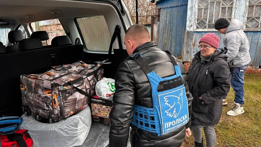 Мужчина в синем жилете с изображением голубя мира стоит в открытом багажнике автомобиля в окружении гуманитарной помощи, а женщина и мальчик наблюдают за ним.