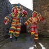 Dois caretos, figuras mascaradas com trajes coloridos, se apresentam em um beco estreito de pedra em Podence, Portugal.