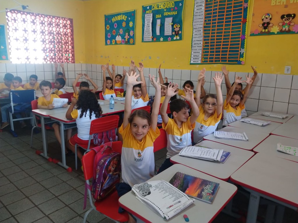 Em uma sala de aula brasileira, os alunos levantam as mãos com entusiasmo.