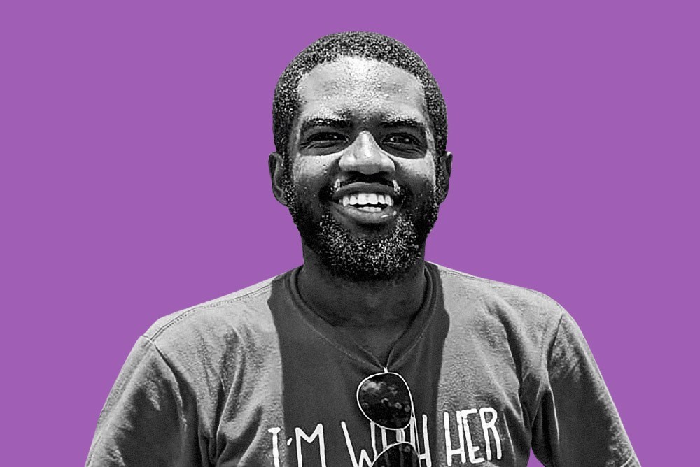Henry Jevanic smiles in a black and white portrait with a purple background, wearing a t-shirt with the slogan 'I'm with her' and sunglasses.