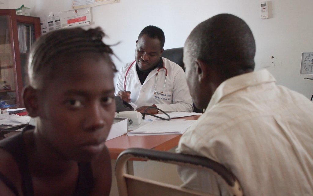 Un médico atiende a un paciente en una clínica de Mozambique, con otro paciente visible en primer plano.