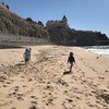 Crianças caminham em uma praia de areia perto de um penhasco com um prédio no topo, com ondas do oceano em um dia ensolarado.