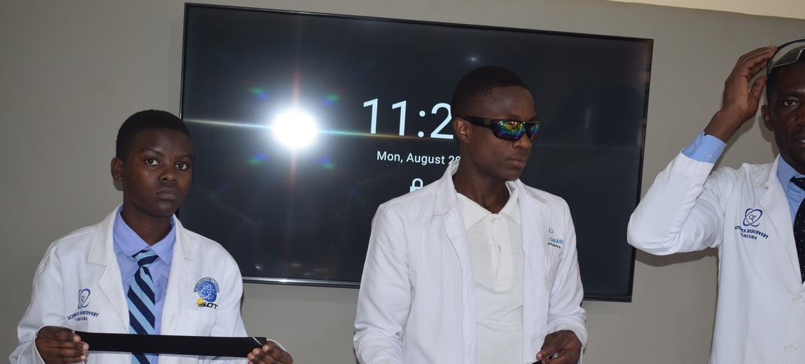 Wanafunzi watatu vijana wa kiume Weusi waliovaa makoti meupe ya maabara na miwani ya usalama wanafanya jaribio la sayansi mezani wakiwa na vifaa vya maabara. Skrini kubwa inayoonyesha muda iko nyuma.