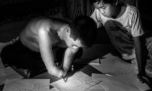 A boy in Viet Nam looks on as his father, whose lower arms were amputated sketches out hands on pieces of paper.