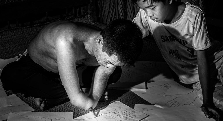 A boy in Viet Nam looks on as his father, whose lower arms were amputated sketches out hands on pieces of paper.