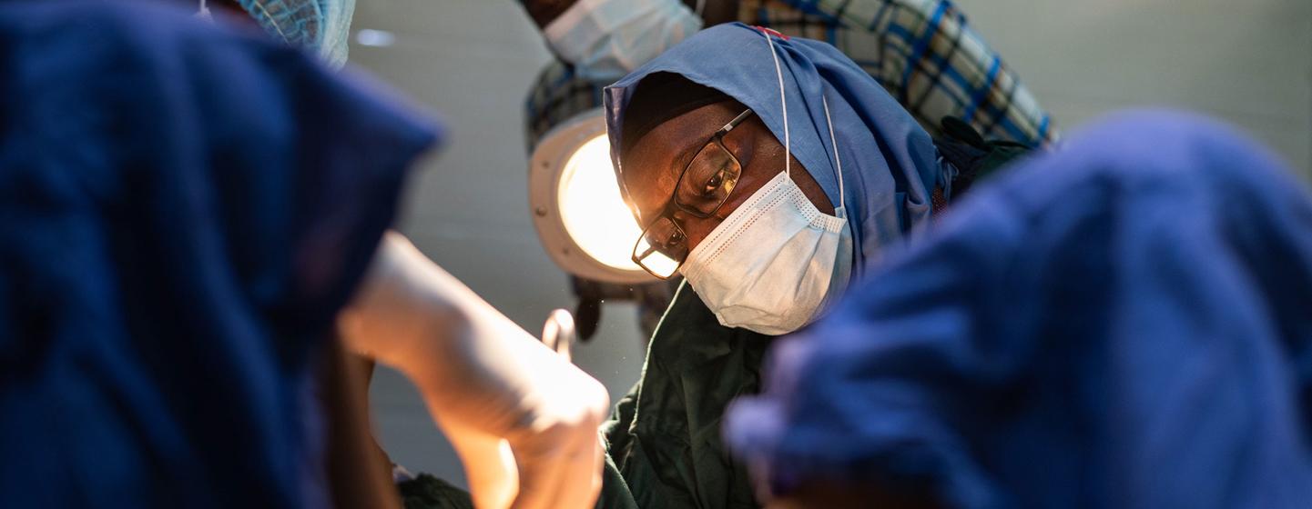 Une médecin concentrée, portant des lunettes et un masque, réalise une intervention médicale, entourée de son équipe dans une salle d'opération.