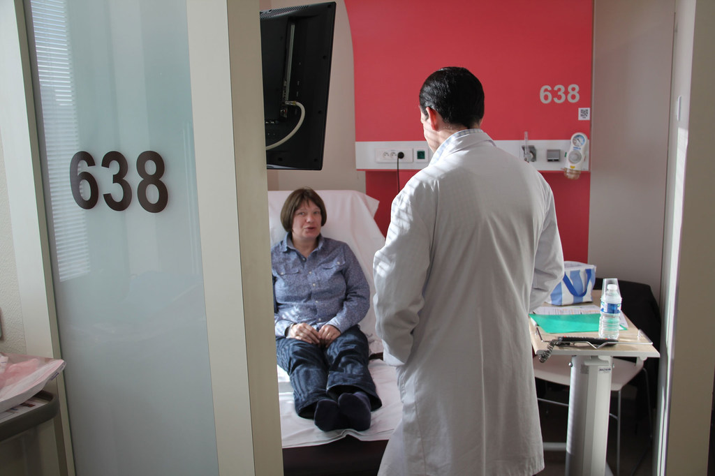 Un médico varón con bata blanca atiende a una paciente sentada en una camilla de exploración en una habitación de hospital, identificada como el Centro Léon Bérard en Lyon, Francia.