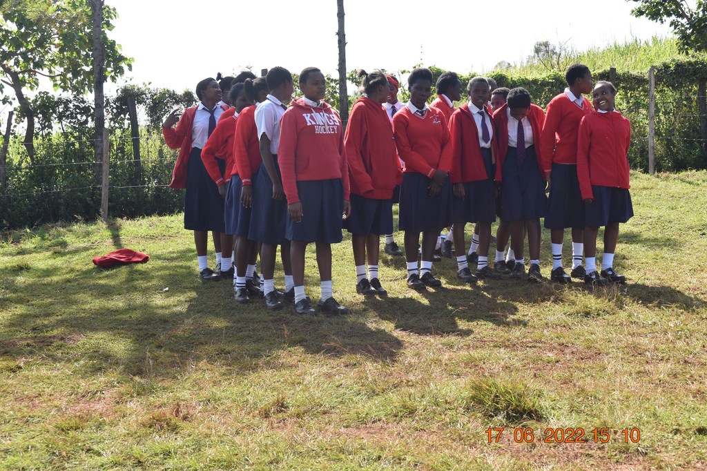 Kundi la wanafunzi wa kike waliovalia sweta nyekundu na sketi za bluu wakiwa wamesimama kwenye uwanja wenye nyasi katika shule ya Kakenya huko Narok, Kenya.