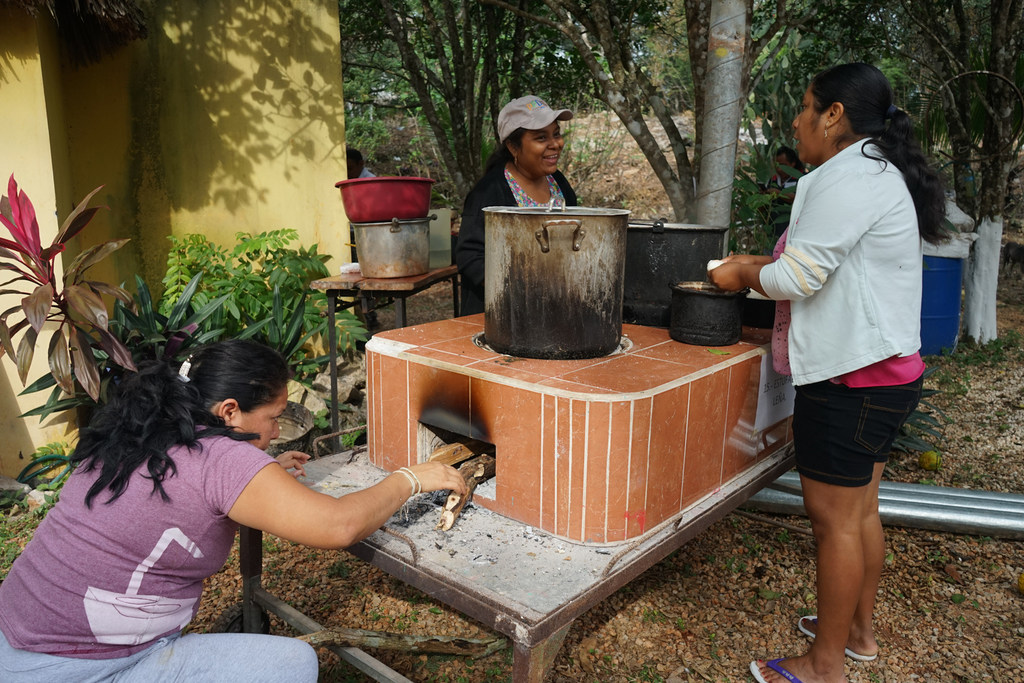 Mujer metiendo leña en un horno con el que otras dos cocinan