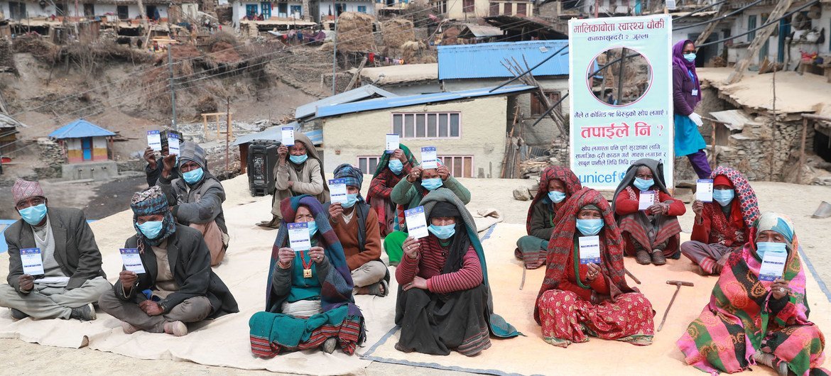 Elderly people at a remote village in Nepal hold up their vaccination cards after receiving COVID-19 vaccines. A sign behind them encourages everyone to receive the vaccines and highlights ways to protect oneself from the disease.