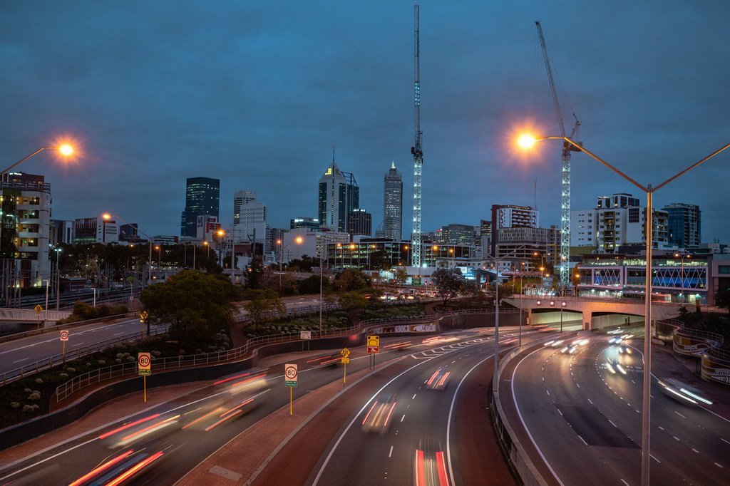 Fotografía de larga exposición de una autopista en Perth, Australia, al atardecer, con estelas de luz de los coches en movimiento y el perfil urbano de la ciudad al fondo.
