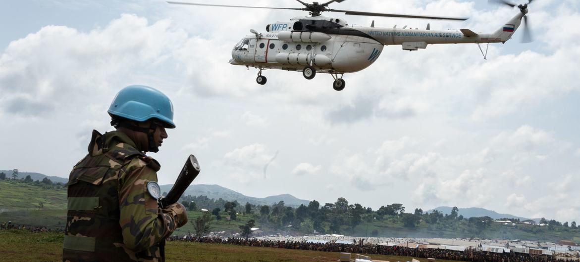 A UN blue helmet from the MONUSCO peacekeeping mission stands guard as a UN helicopter delivers aid and humanitarian personnel to Rhoe IDP camp, Ituri, Democratic Republic of the Congo.