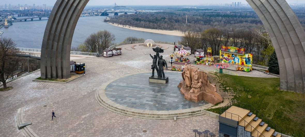 Вид сверху на арку Независимости в Киеве на реку Днепр на заднем плане. Под аркой видны детская площадка и карусель, вокруг очень мало людей.
