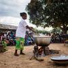 Mwanamke aliyevaa fulana nyeupe na sketi yenye muundo wa kijani akipika kwenye sufuria kubwa ya chuma juu ya moto wazi nje, huku kundi la watu likitazama.