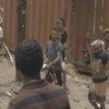 يقف عدد من الرجال المسلحين في منطقة متربة، وخلفهم حاوية شحن كبيرة. ويظهر رجل في المقدمة من الخلف، ينظر باتجاه المجموعة.