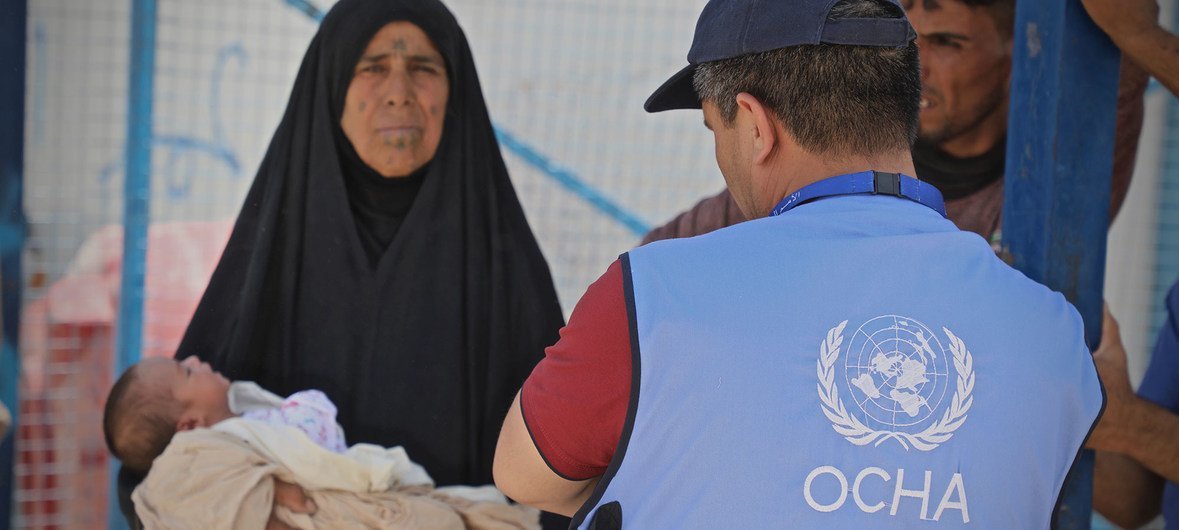 संयुक्त राष्ट्र के मानवीय सहायता कर्मचारी अल होल शिविर (सीरिया) में एक स्वास्थ्य क्लीनिक में एक महिला की मदद करते हुए जो अपने चार दिन के पोते को वहाँ लेकर आई थी. (16 जून 2019) 