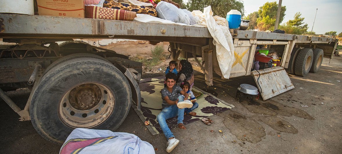 On 11 October 2019 in the Syrian Arab Republic, a woman and children sit underneath a truck as people displaced from Ras al-Ain arrive in Tal Tamer, having fled escalating violence.