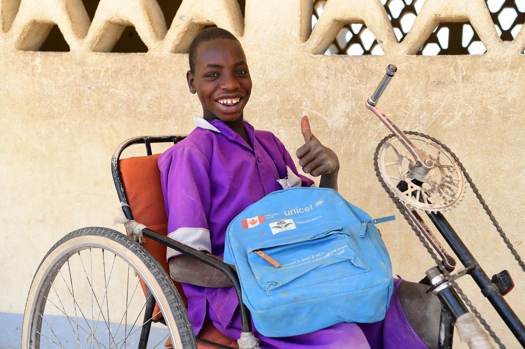 Foto de un niño de raza negra en silla de ruedas, saluda a cámara con el pulgar hacia arriba mientras sonríe y sujeta una mochila azul de Unicef