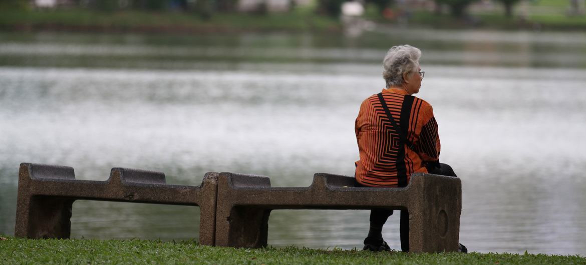 An elderly woman sits alone on a bench near a pond in Thailand.