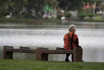 An elderly woman sits alone on a bench near a pond in Thailand.