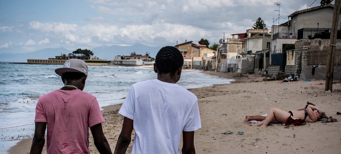 Dois irmãos adolescentes da Gâmbia que viajaram sem os pais pelo mar Mediterrâneo caminham por uma praia na Itália.