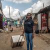 A Kenyan woman stands on a community street pulling a cooler on a trolley. She is wearing a black polo shirt and blue jeans, with a few people and buildings visible in the background.