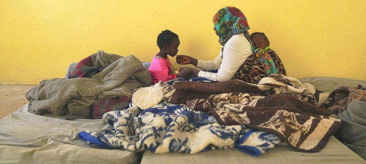 أم محتجزة مع طفليها أحدهما نائم على ظهرها والأخر يتناول الخبز من يدها في أحد مراكز الاحتجاز في بنغازي، ليبيا