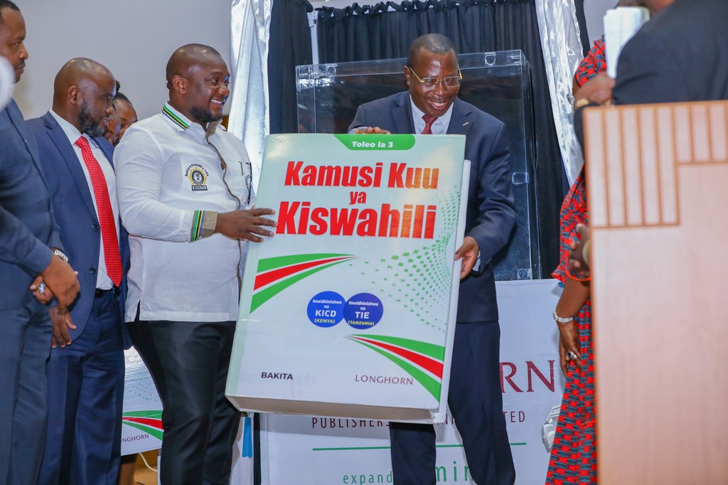 Mwanamume aliyevaa suti na mwanamume aliyevaa shati jeupe akiwa ameshika nakala kubwa ya kitabu cha 'Kamusi Kuu ya Kiswahili' katika hafla.