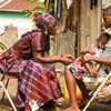 Wanawake wawili na mtoto mmoja wameketi kwenye viti vya nje. Mwanamke mzee aliyevaa gauni la rangi na kitambaa cha kichwani anaegemea mwanamke mdogo aliyemshika mtoto aliyelala. Mwanamke mdogo, akiwa amevaa gauni lenye muundo, anamtazama mwanamke mzee huku akimbembeleza mtoto.