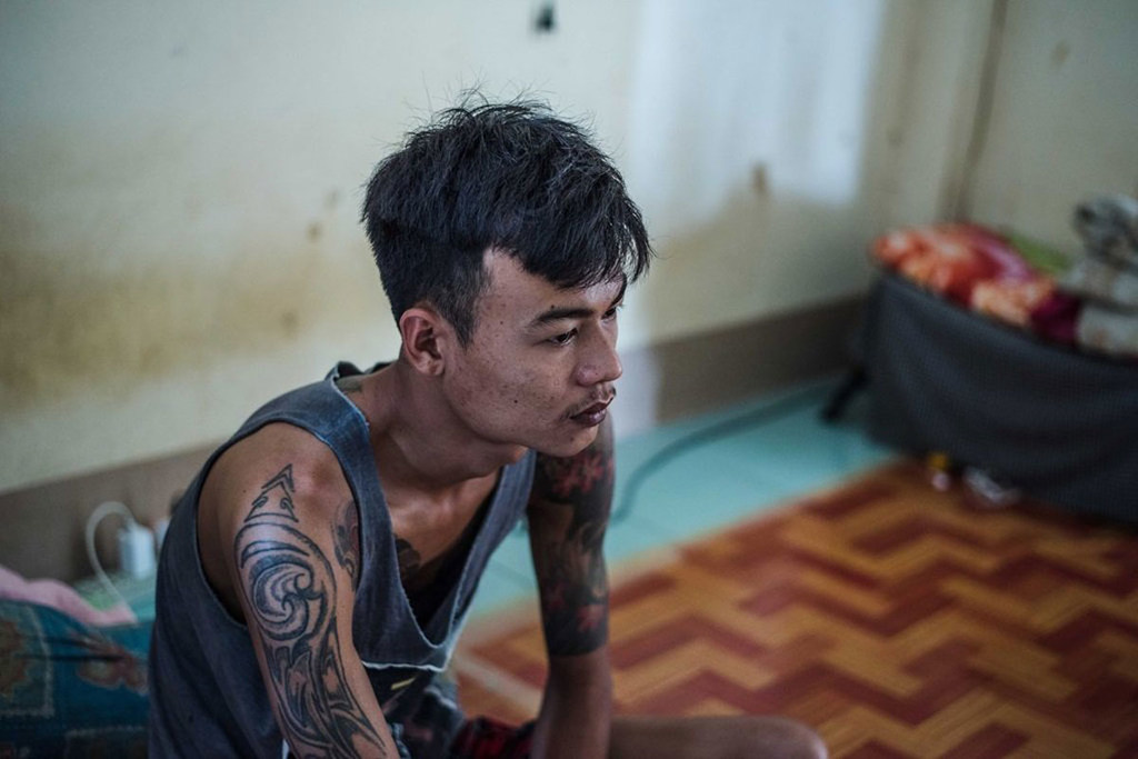 Un hombre asiático tatuado está sentado en el suelo de una habitación, mirando hacia otro lado con expresión sombría.