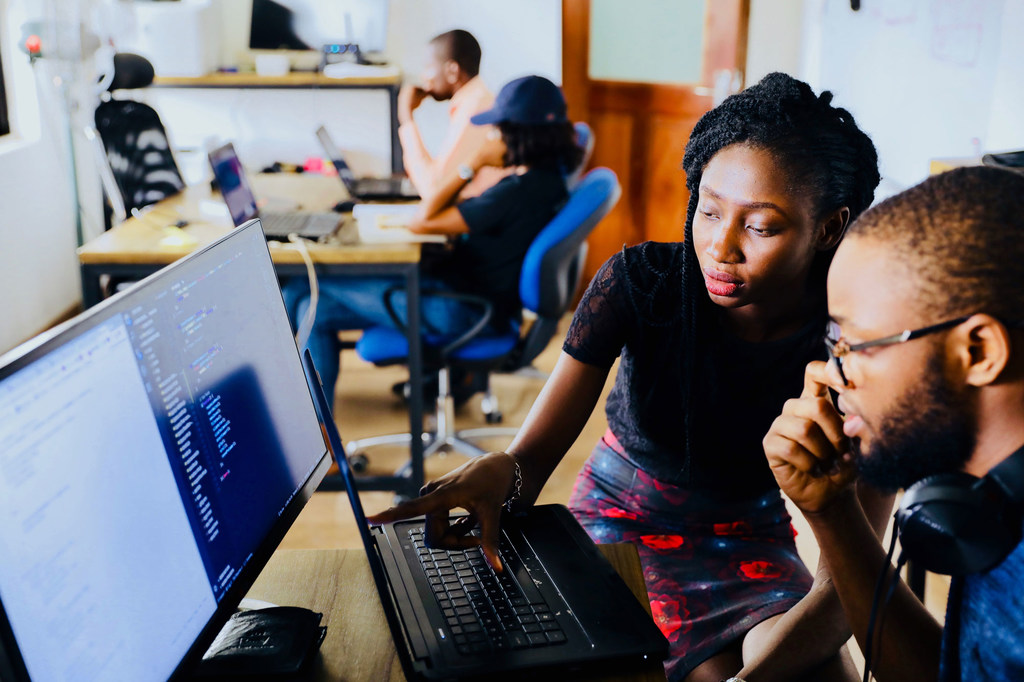 Uma mulher e um homem afro-americanos colaboram em um código exibido em um laptop e um monitor em um ambiente de escritório.