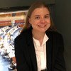 Портрет улыбающейся женщины в черном пиджаке и белой рубашке, стоящей перед окном, выходящим в большой конференц-зал.