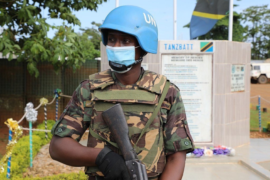 Mlinda amani wa Tanzania aliyevaa kofia ya chuma MONUSCO na sare ya kujificha amesimama mbele ya bamba la ukumbusho lenye bendera ya Tanzania.