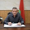 Валентин Рыбаков сидит за столом, подписывая документы, на фоне белорусского флага.